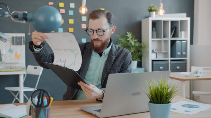 Smart guy is reading graphs and charts on paper and looking at computer screen in office doing research in workplace. People, technology and information concept.