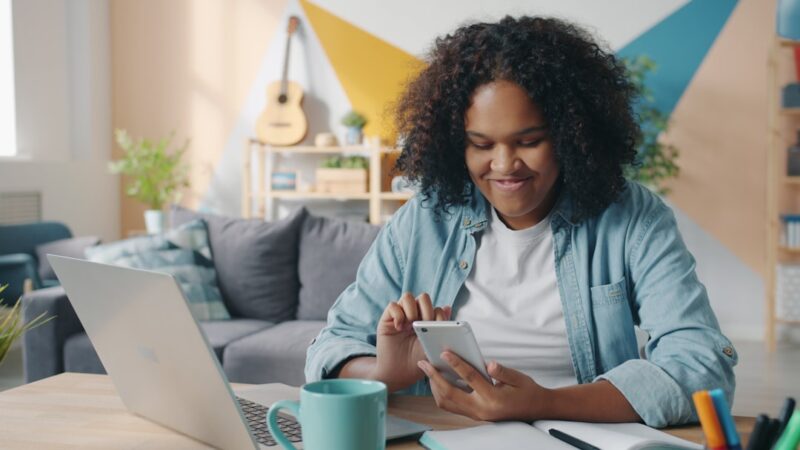 Pretty Afro-American woman student is using smartphone at home at desk smiling touching screen enjoying communication. People and lifestyle concept.